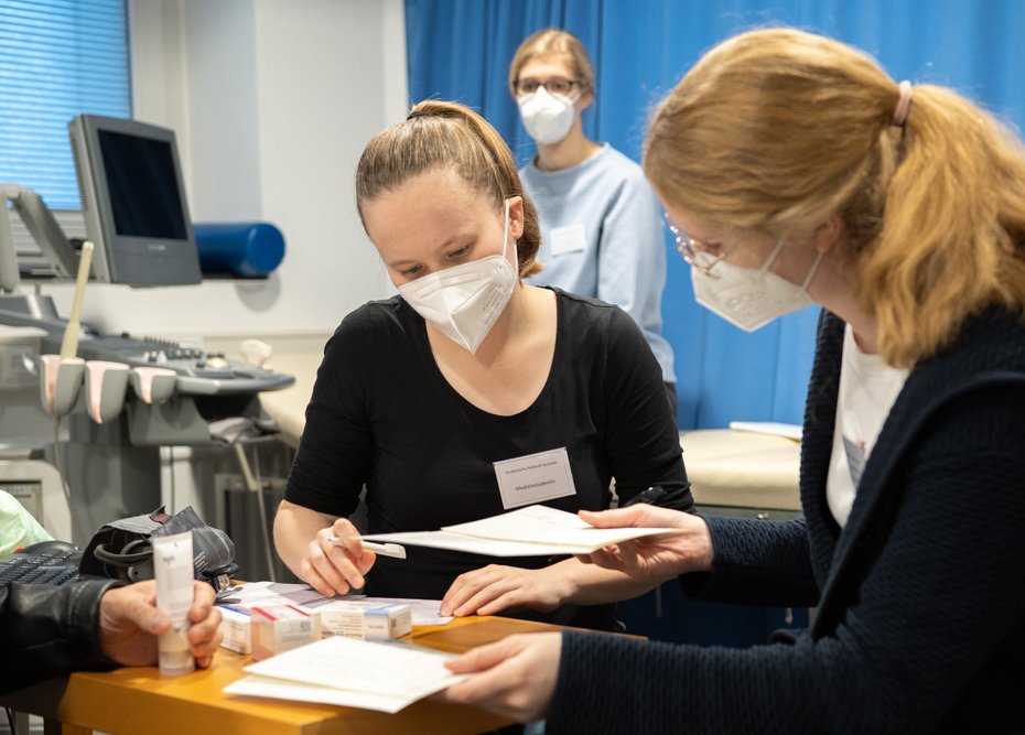 Zwei Studentinnen beraten im Vordergrund einen Patienten, im Hintergrund sitzt eine Ärztin auf einer Behandlungsliege.