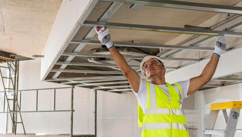 Ein Handwerker in Sicherheitsweste steht mit erhobenen Armen unter einer Deckenkonstruktion.