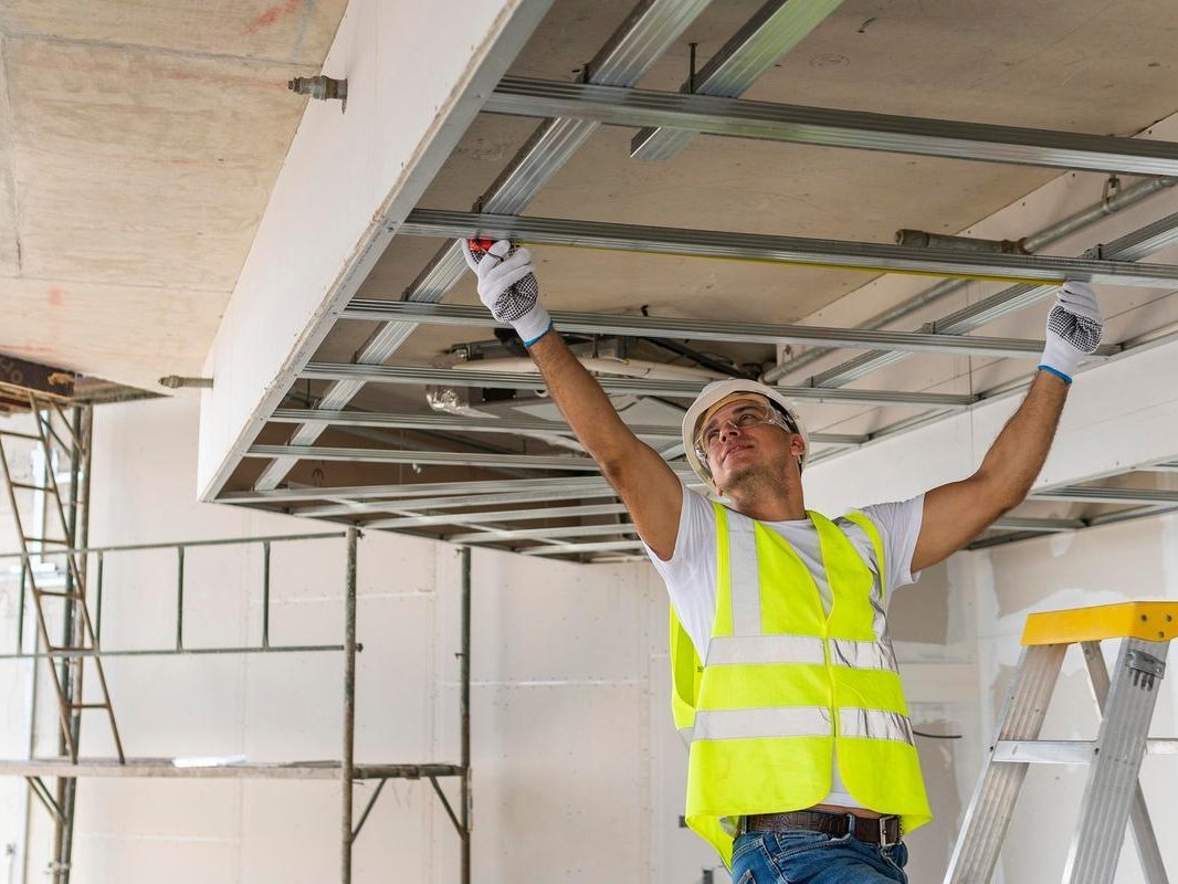 Ein Handwerker in Sicherheitsweste steht mit erhobenen Armen unter einer Deckenkonstruktion.