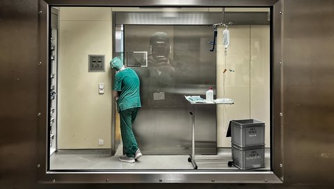 A person in a green coat stands at a metal sliding door to an operating theater and looks in through a small gap.