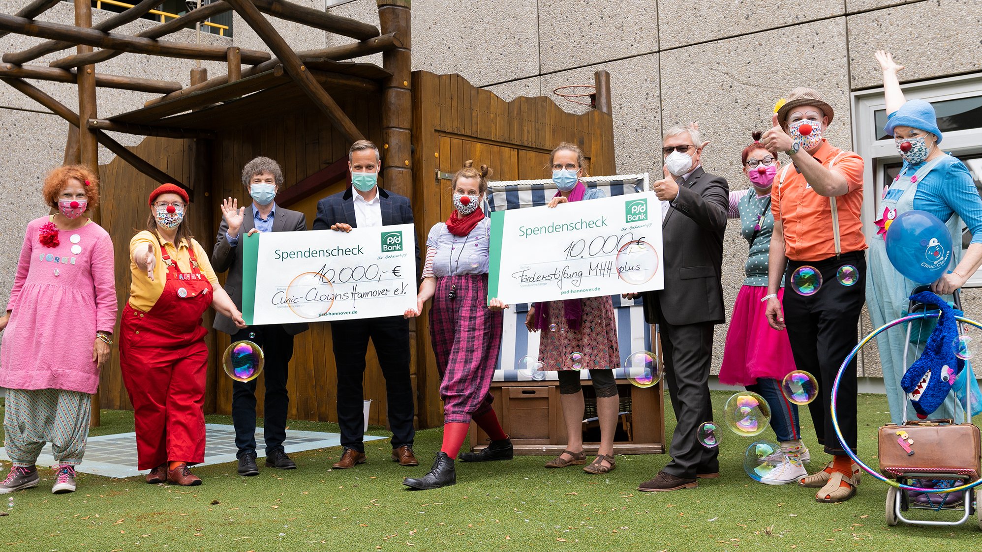 Sechs Clinic Clowns, eine Mitarbeiterin der Kinderklinik, Dr. Eckhard Schenke von MHH plus sowie Torsten Krieger und Holger Hammer von der PSD-Bank halten zwei Spendenschecks in die Höhe. Im Vordergrund sind Seifenblasen zu sehen.