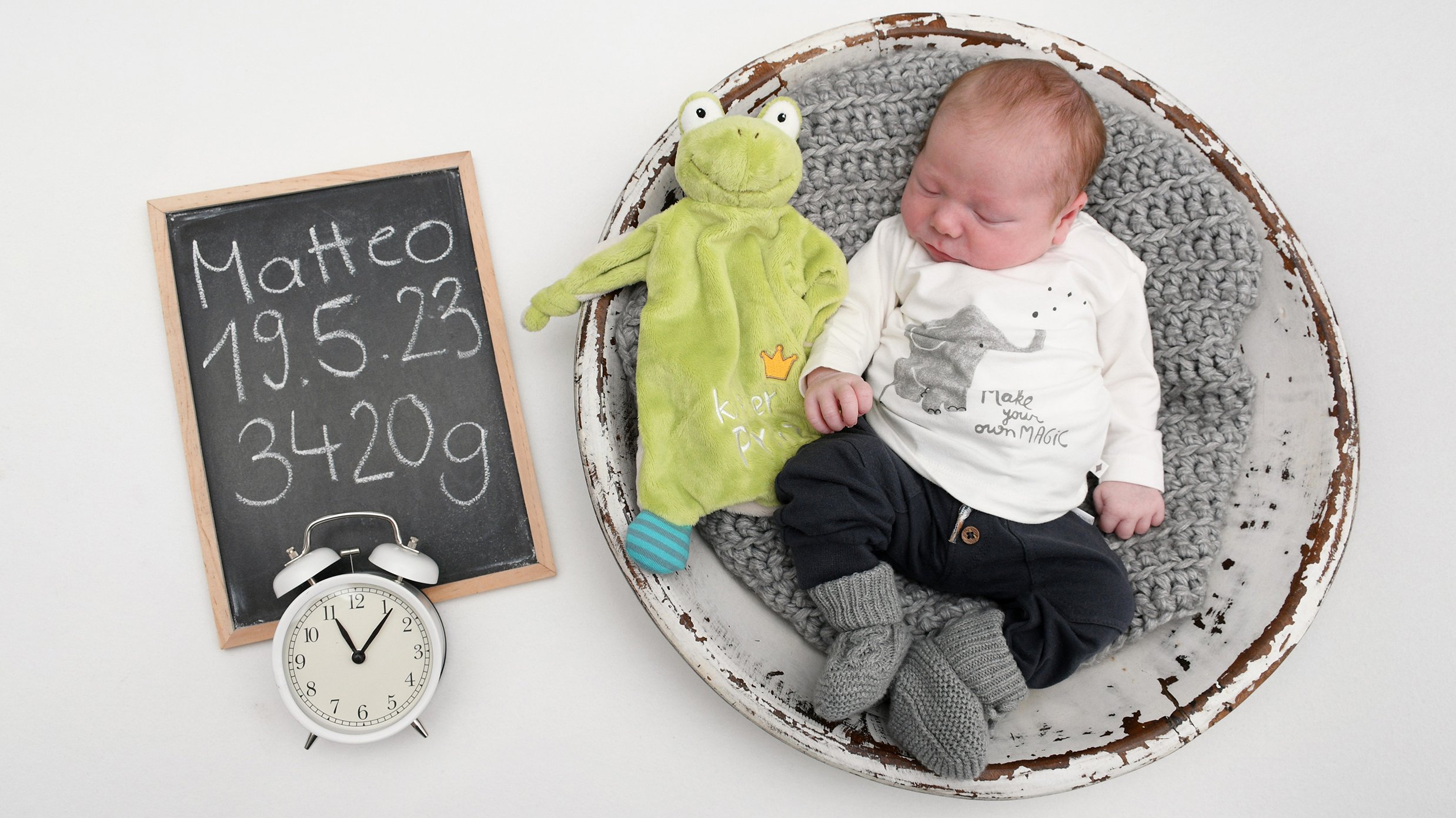 Ein Baby liegt in einem geflochtenen Korb, neben ihm eine Frosch als Stofftier. Neben dem Korb steht eine kleine Kreidetafel mit dem Schriftzug Matteo 19.5.23 3420g sowie ein Wecker.