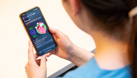 Blick über die Schulter einer jungen Frau mit dunklen Haaren hinunter auf ein Smartphone, das einen Ausschnitt aus der App zeigt.