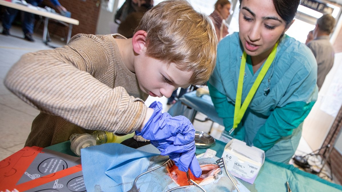 A boy is performing an operation on a stuffed sheep at the OTA stand. An OTA is standing next to him. 