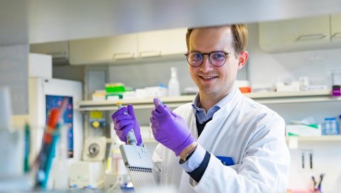 Dr. Bernd Heinrich steht in einem Labor und hält ein Mikropipettiergerät in der Hand.