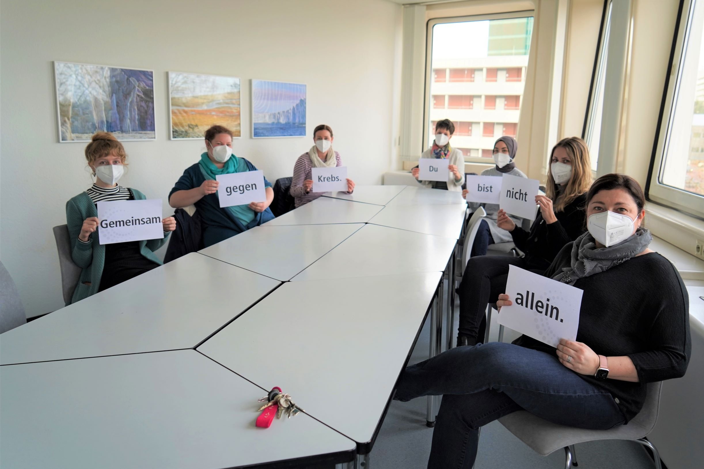 Mitarbeiterinnen vom Team der Psychoonkologie sitzen im Besprechungraum und halten Schilder, auf denen steht "Gemeinsam gegen Krebs. Du bist nicht allein."