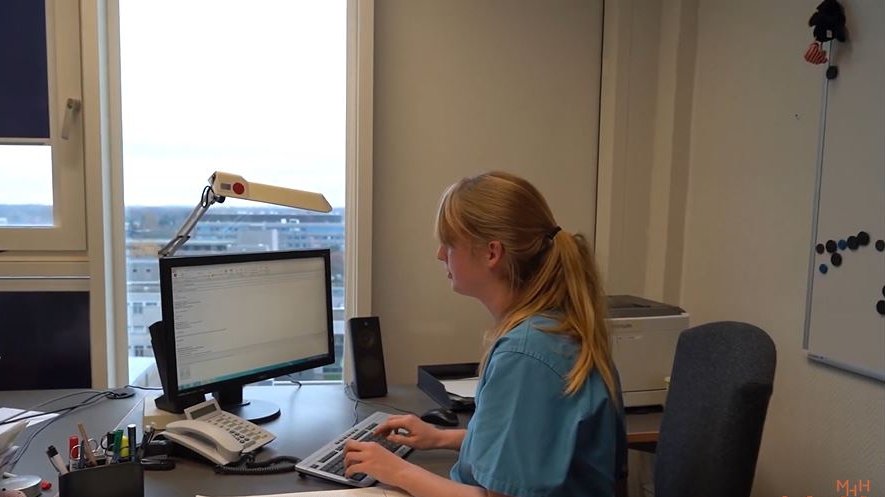 Eine Pflegekraft im blauen Kasack sitzt an einem Schreibtisch und tippt auf einer PC-Tastatur.