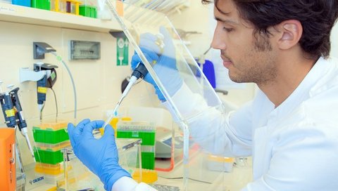 A man in a white coat sits in front of a sterile laboratory bench holding a pipette in his hands.