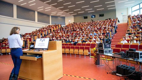 View from the lectern into a packed lecture hall. On the left stands MHH President Denise Hilfiker-Kleiner, addressing the first-year students.