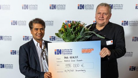 Zwei Männer halten einen Blumenstrauß und symbolischen Förderbescheid in den Händen.