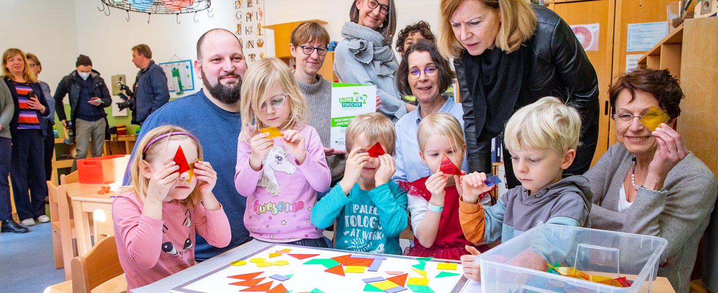 Mehrere Erwachsene und Kinder stehen an einem Tisch mit einem Brettspiel: links Kitaleiter Martin Fulst, stehend Dr. Andrea Handke, Region Hannover und Martina Saurin, MHH-Vizepräsidentin, rechts. 