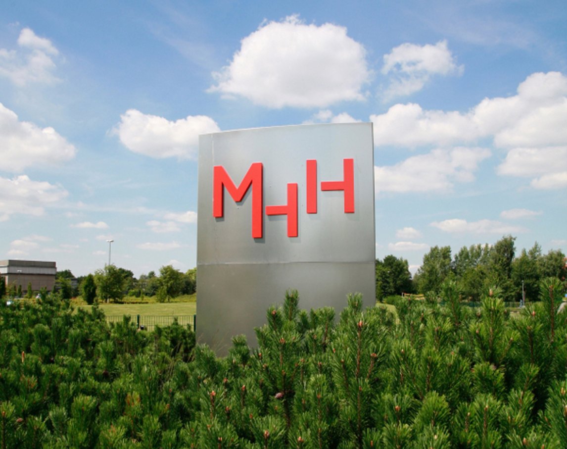 Edelstahlstehle mit rotem MHH-Logo, im Vordergrund Kiefernhecke, im Hintergrund blauer Himmel mit Wolken