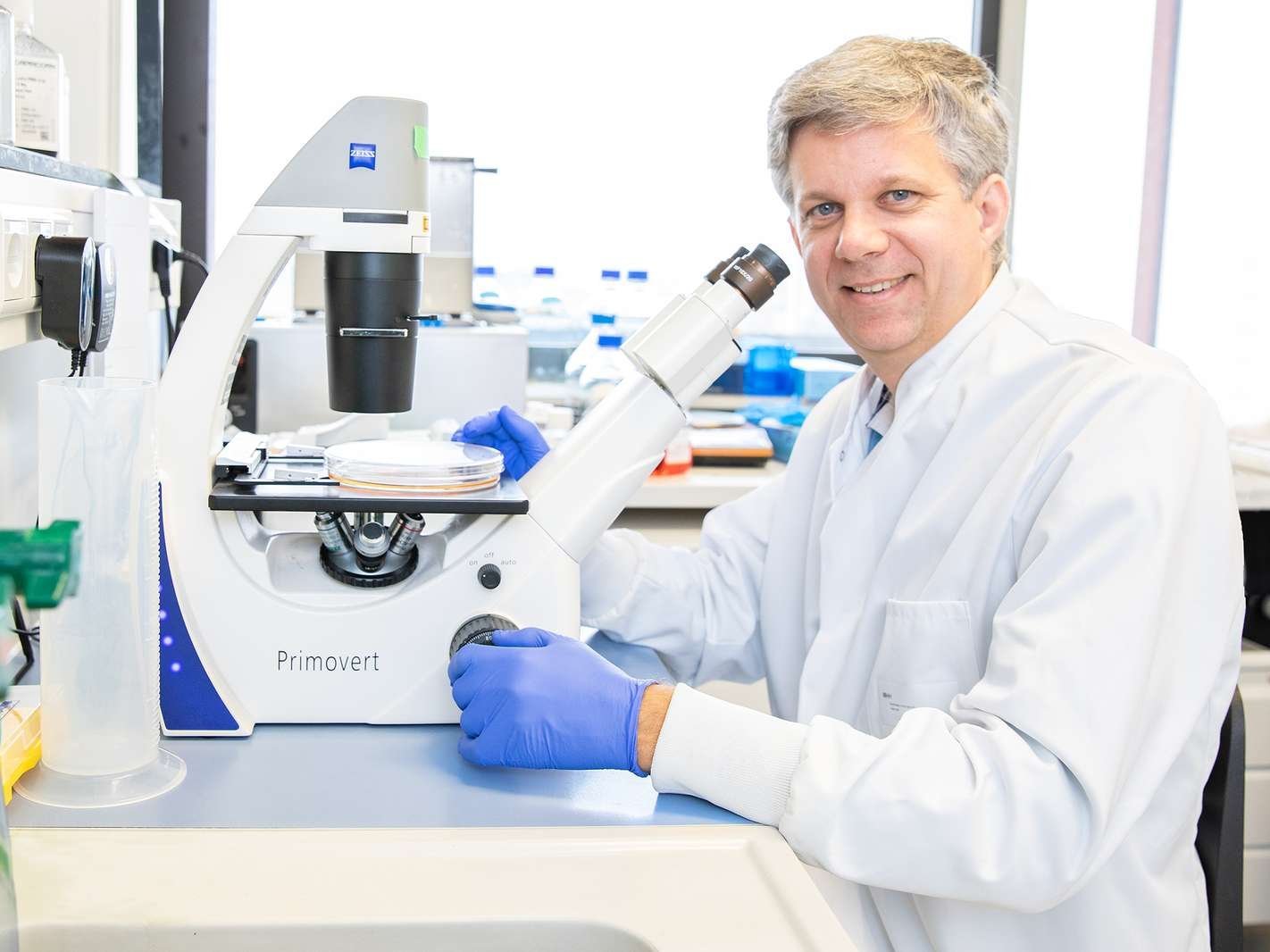The photo shows Prof. Dölken in a white lab coat. He is sitting at a bench in a laboratory with a microscope in front of him. He is looking into the camera.