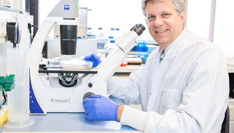 The photo shows Prof. Dölken in a white lab coat. He is sitting at a bench in a laboratory with a microscope in front of him. He is looking into the camera.