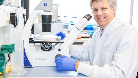Auf dem Foto ist Prof. Dölken in einem weißen Laborkittel zu sehen. Er sitzt in einem Labor an einer Laborbank und hat ein Mikroskop vor sich stehen. Er blickt in die Kamera.