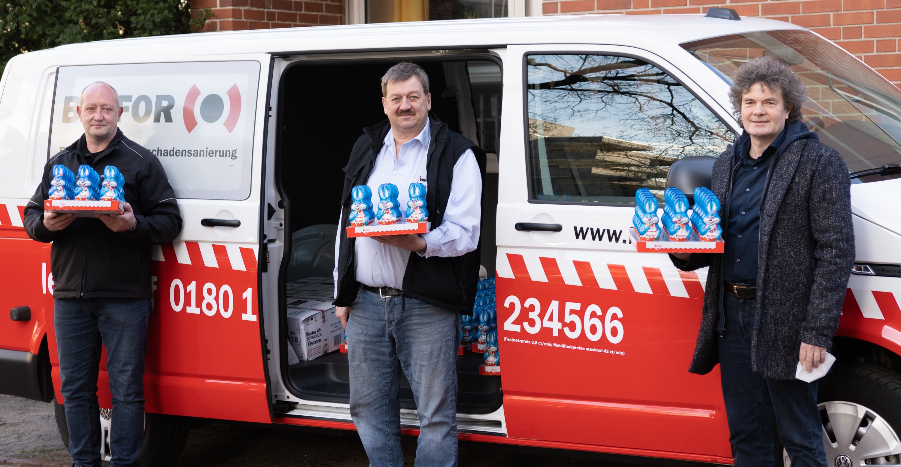 Zwei Mitarbeiter der Firma Belfor stehen mit Dr. Schenke für einem Lieferwagen der Firma. Alle drei haben eine kleine Papppalette mit Schoko-Osterhasen in der Hand. 