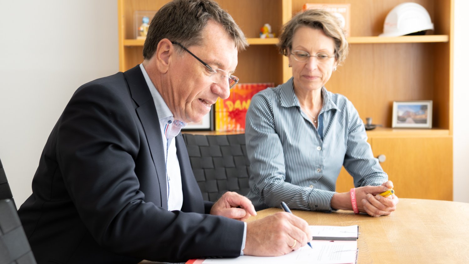 Herr Fischer unterschreibt im Vordergrund, Frau Saurin sitzt daneben und schaut zu.