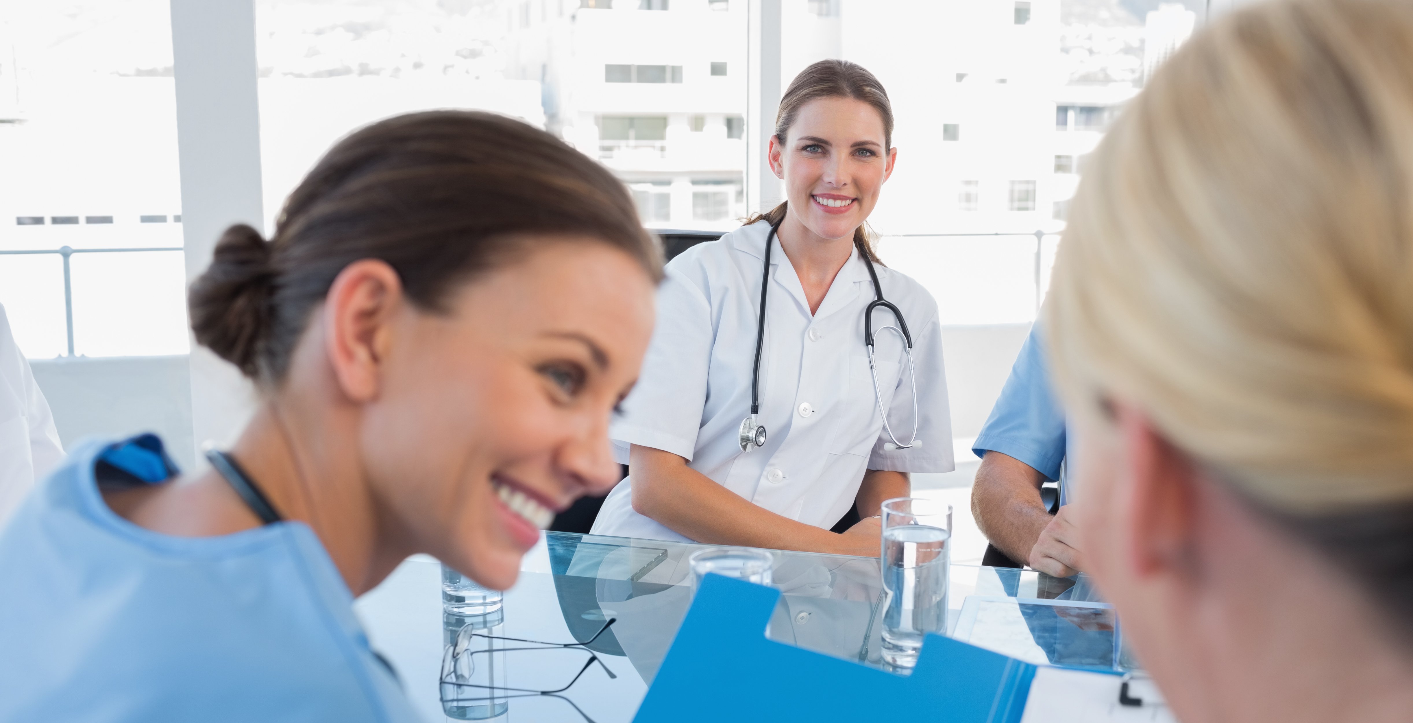 Das Bild zeigt drei Frauen an einem Besprechungstisch, sie tragen einen Kasak bzw. weißen Kittel jeweilsmit Stetoskop. Eine Frau ist von hinten zusehen, dien anderen beiden lächeln.