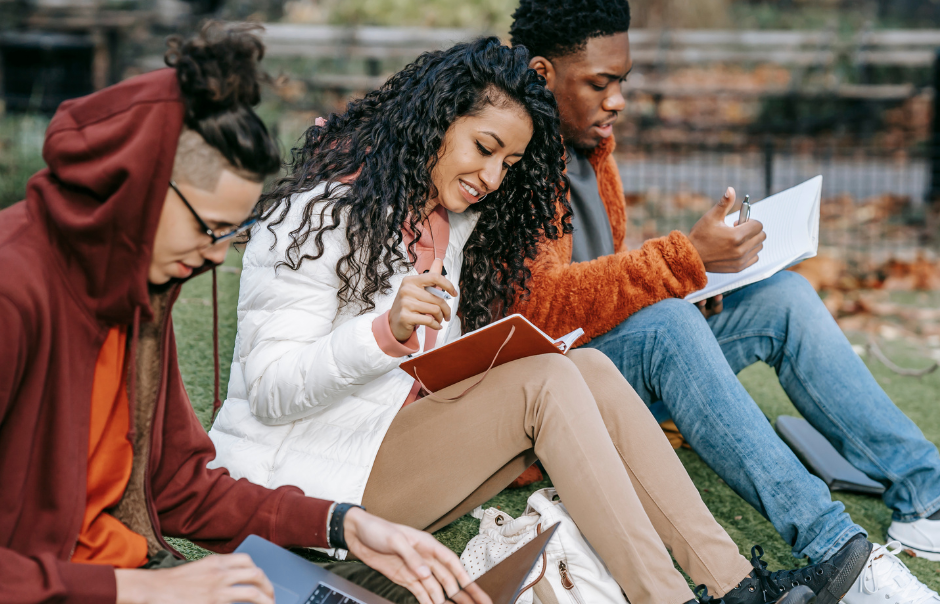 Drei junge Menschen sitzen nebeneinander auf einer Rasenfläche und lesen und schauen auf einen Laptop. 