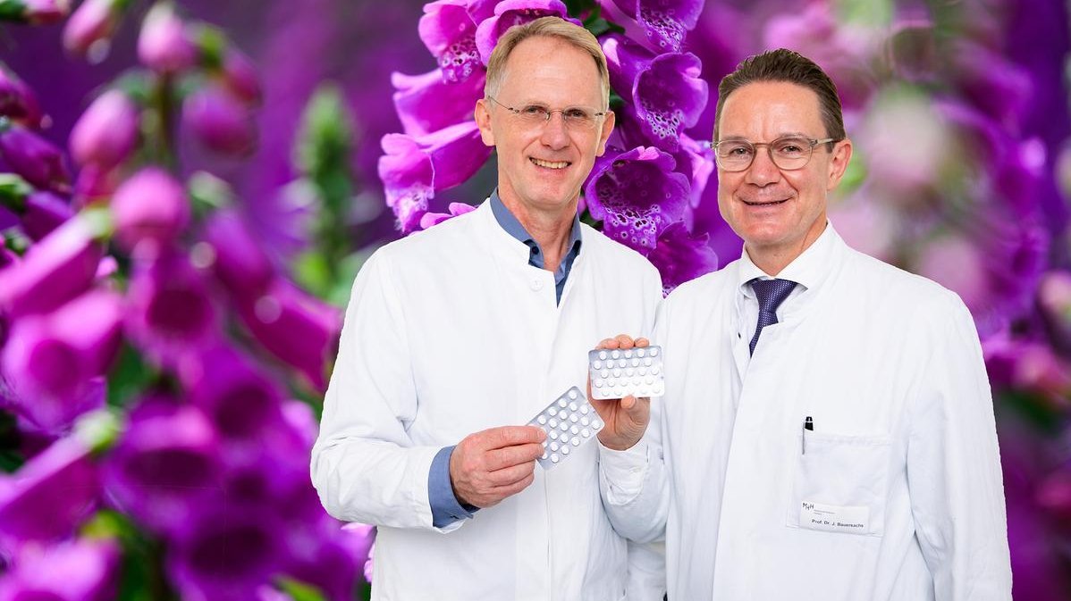 Die Kardiologen Professor Dr. Udo Bavendiek (links) und Professor Dr. Johann Bauersachs stehen nebeneinander und halten Tablettenpackungen in den Händen. Im Hintergrund ein Bild mit der Pflanze Fingerhut.  