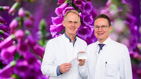 Zwei Männer im weißen Kittel stehen vor einem Foto mit rotem Fingerhut und halten Tabletten-Blister in der Hand.