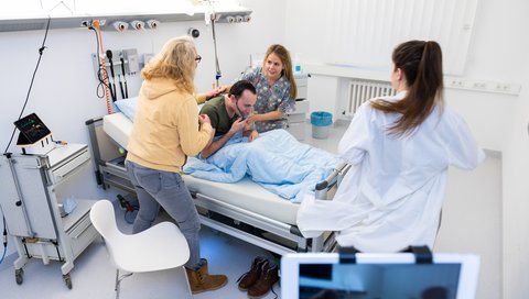 Studierende und Pflegeauszubildende stehen um ein Krankenbett, in dem ein Patient mit Atemmaske liegt und üben eine Notfallsituation.