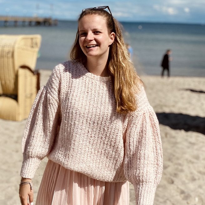 Deborah Meybohm steht an einem Strand, im Hintergrund sieht man Strandkörbe und das Meer. 