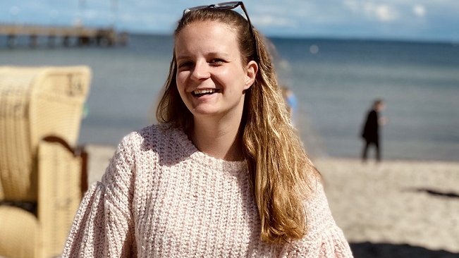 Deborah Meybohm steht an einem Strand, im Hintergrund ist eine Strandkorb und das Meer zu sehen. 