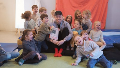 Kinder der MHH-Kita sitzen um einen Mann mit Buch auf Matten auf dem Boden. 