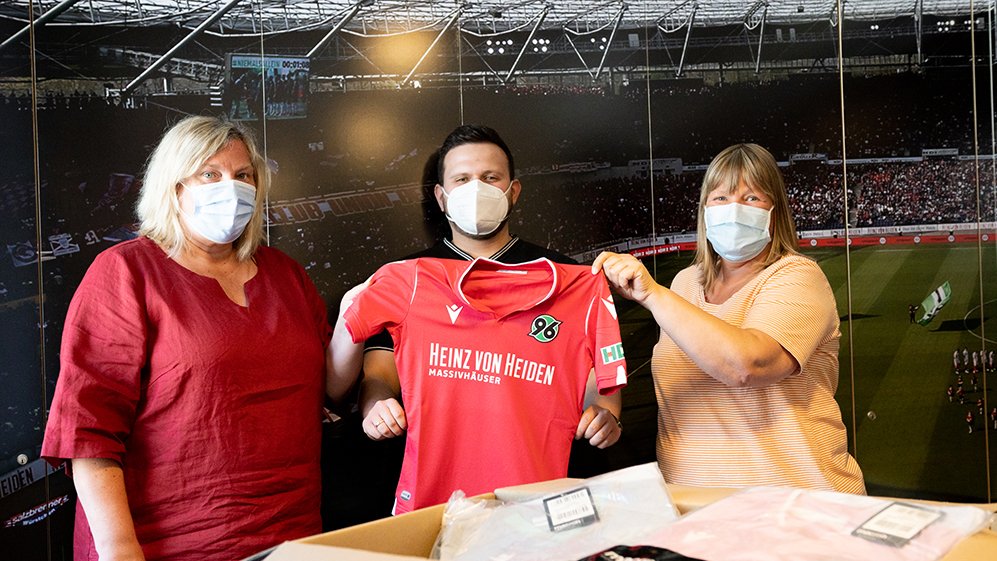 Claudia Erzfeld-Dokanikis und Annette Wiens von der Spieloase halten gemeinsam mit Juri Sladkov von der Initiative 96plus ein rotes Hannover96-Trikot hoch.