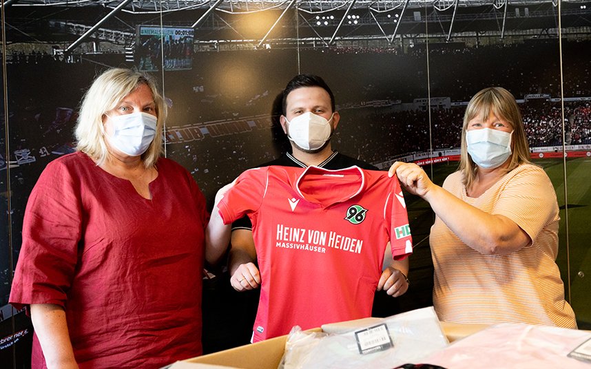 Claudia Erzfeld-Dokanikis und Annette Wiens von der Spieloase halten gemeinsam mit Juri Sladkov von der Initiative 96plus ein rotes Hannover96-Trikot hoch.