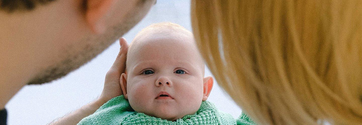 zwischen den angeschnittenen Hinterköpfen eines Vaters und einer Mutter blickt ein Baby den Betrachter direkt an.