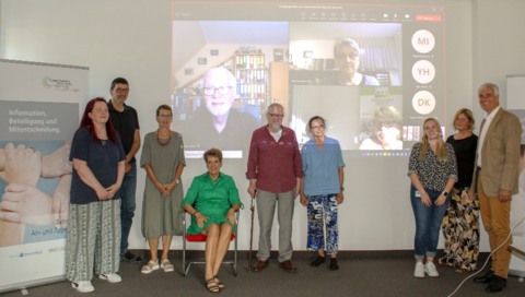 Mehrere Mitglieder des CCC-Patientenbeirats stehen oder sitzen vor einer weißen Wand, an der per Videokonferenz zugeschaltete weitere Mitglieder zu sehen sind. 