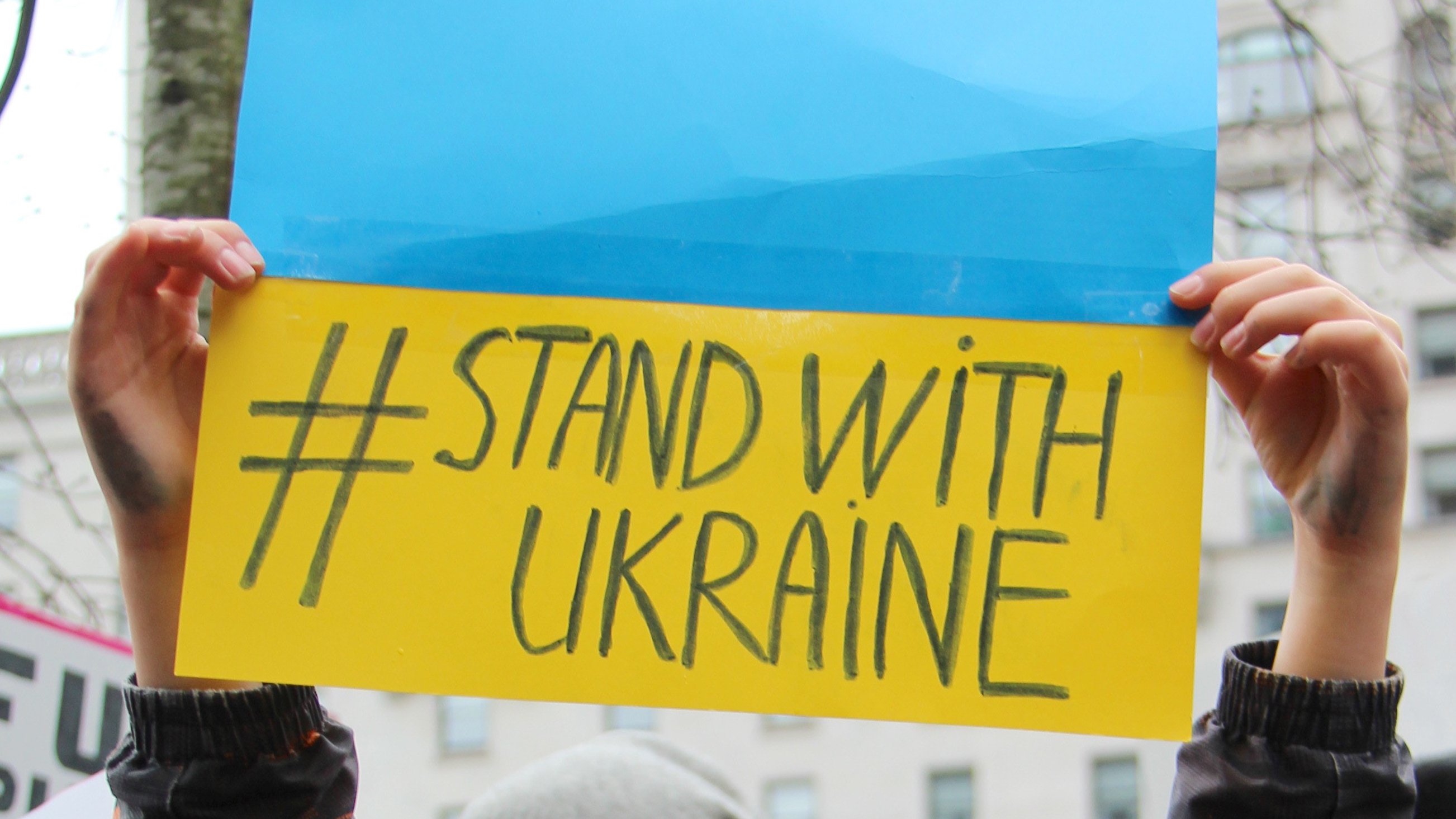 Eine Person hält ein Papier mit den Farben der Urkraine-Flagge Blau und Gelb hoch. Auf dem Papier steht #stand with Ukraine.
