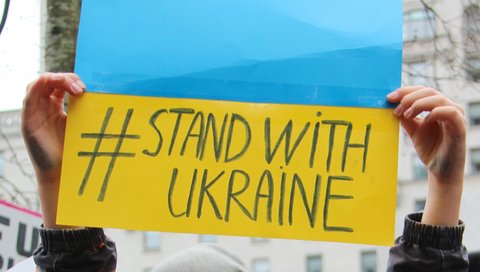 Eine Person hält ein Papier mit den Farben der Urkraine-Flagge Blau und Gelb hoch. Auf dem Papier steht #stand with Ukraine.