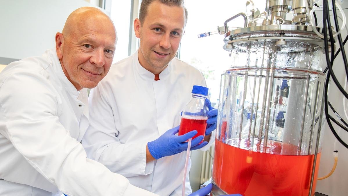 Two researchers in white lab coats are standing in a laboratory in front of a stirred-tank bioreactor. 