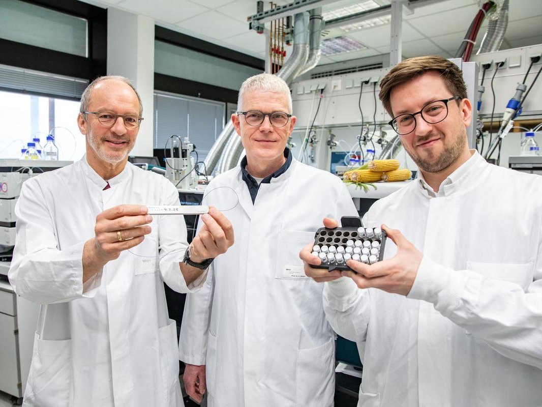 Drei Männer in weißen Kitteln stehen in einem Forschungslabor und betrachten eine Halterung mit Probenmaterial für eine Untersuchung im Massenspektrometer.