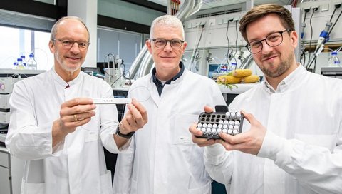 Drei Männer in weißen Kitteln stehen in einem Forschungslabor und betrachten eine Halterung mit Probenmaterial für eine Untersuchung im Massenspektrometer.