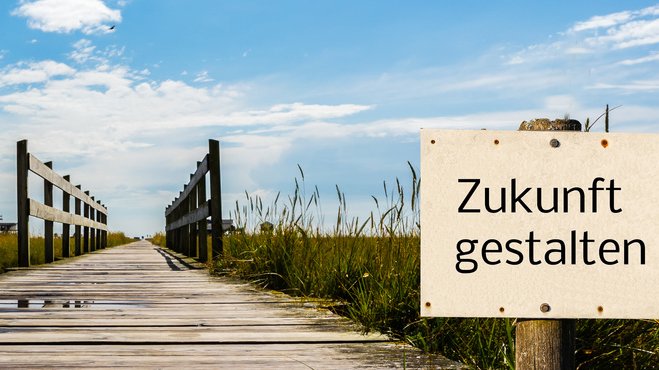 ein Steg, der über eine Wiese führt; am rechten Bildrand steht ein Schild mit der Aufschrift "Zukunft gestalten"