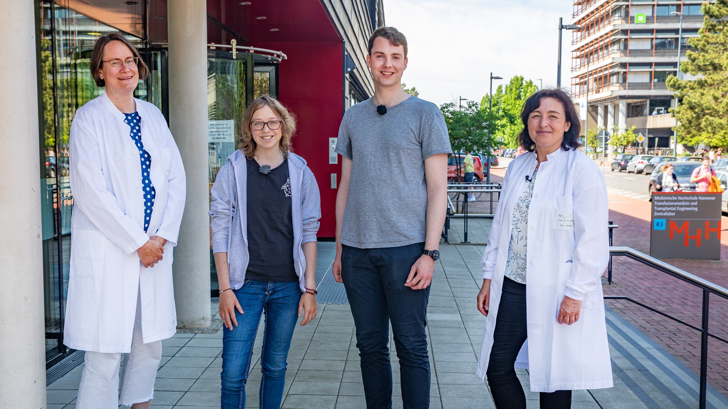 Sarah Schott, Nick Seidel Professorin Dr. Britta Maecker-Kolhoff  und Professorin Dr. Britta Eiz-Vesper stehen vor dem Gebäude der MHH Blutspende und schauen in die Kamera.