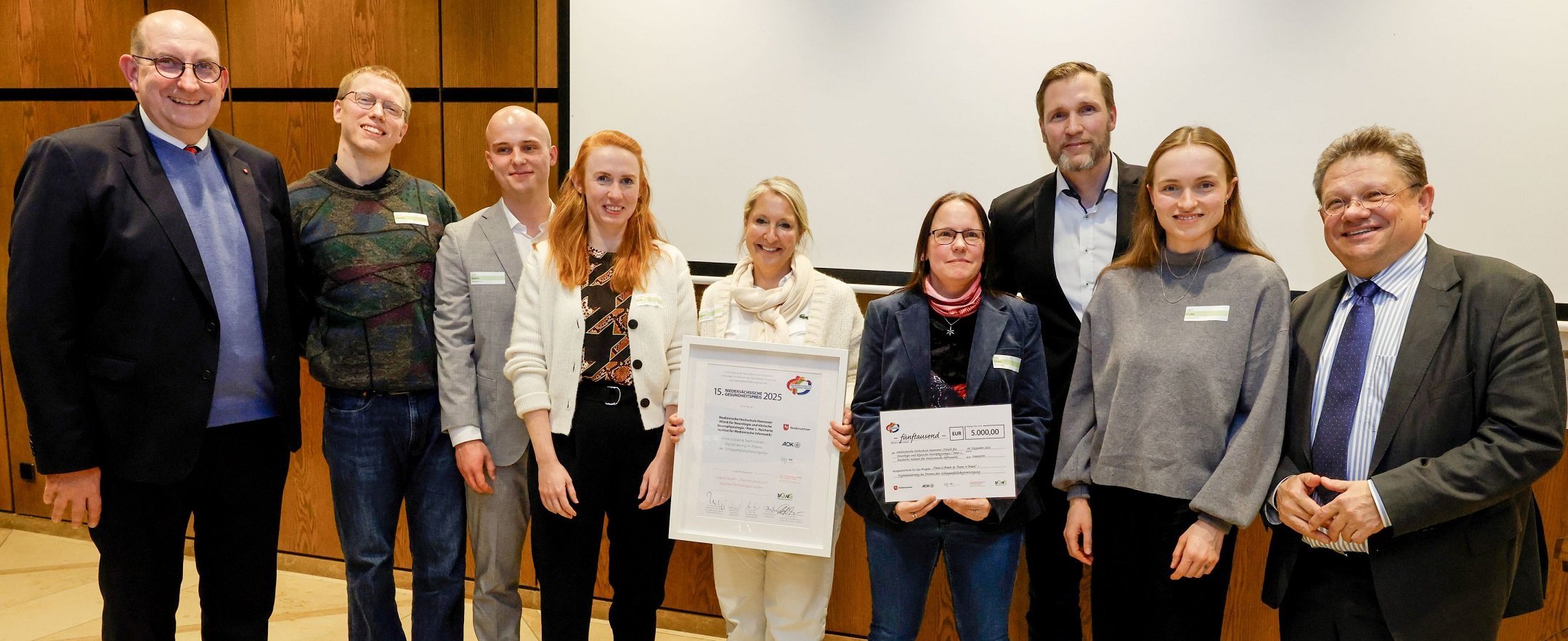 Neun Personen stehen für ein Gruppenfoto nebeneinander, zwei halten Urkunden in den Händen.  