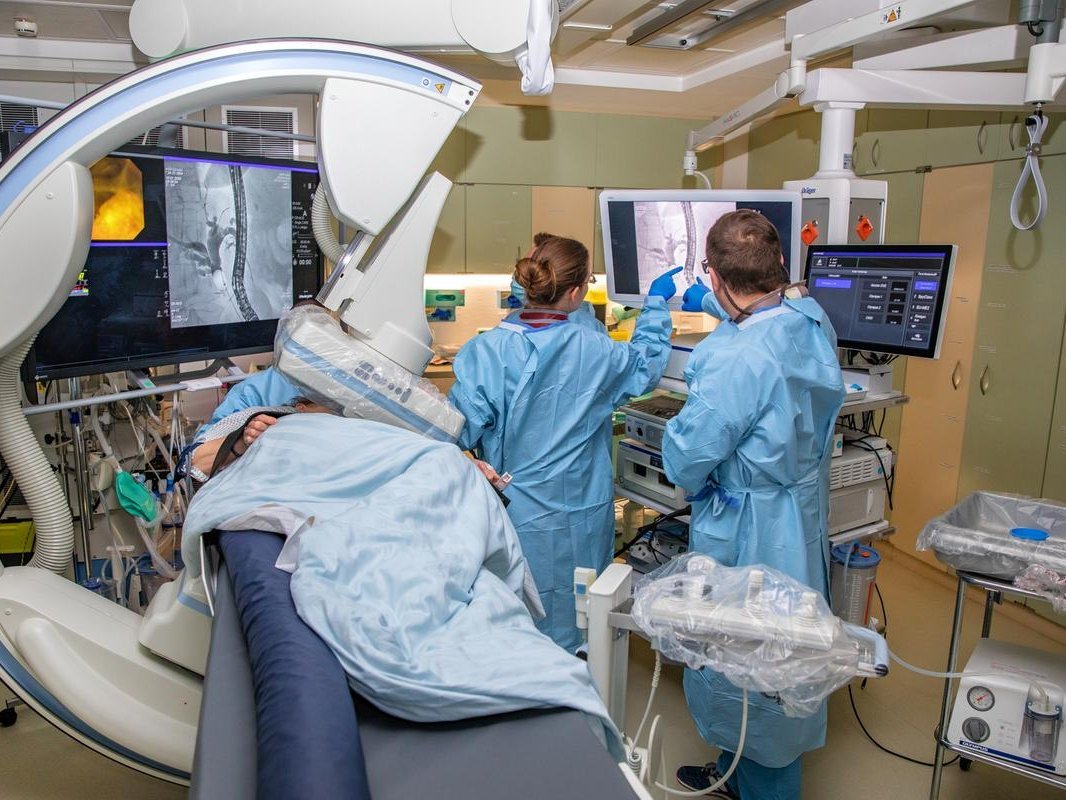 Eine Frau liegt unter einer Apparatur. Rechts von ihr stehen zwei Personen in blauen Kitteln, sie zeigen auf einen Monitor.