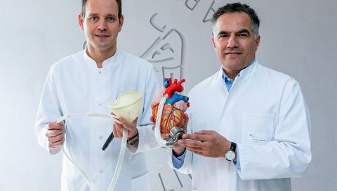 Dr. Schmack hält den Kunststoffbeutel des reBEAT-Systems in der Hand, während Professor Ruhparwar ein Herzmodell mit einem implantierten, herkömmlichen Linksherzunterstützungssystem zeigt.