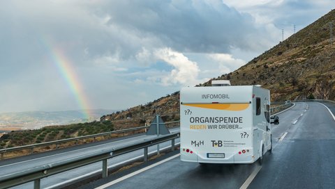 Ein Info-Camper mit der Aufschrift „Organspende: Reden wir drüber“ fährt über eine regennasse Straße; am Horizont zeigt sich ein Regenbogen. 