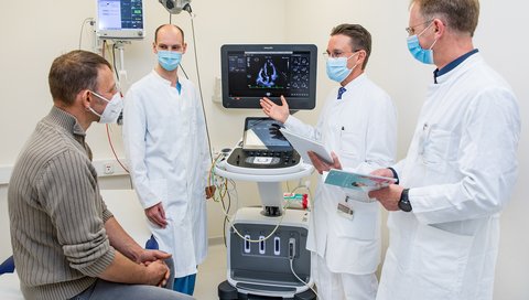 Patient Thorsten S. sitzt auf einer Patientenliege, rechts daneben stehen Dr. Fabian Rathje, Professor Dr. Johann Bauersachs und Professor Dr. Udo Bavendiek. In der Mitte ein Bildschirm mit einer Aufnahme des Herzens.  