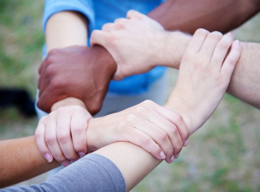 Fünf Hände von Menschen unterschiedlicher Hautfarbe umschlingen jeweils ein anderes Handgelenk und bilden somit einen Kreis.