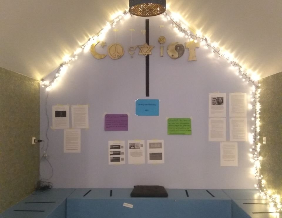 Foto von gestalteter Aktionskabine: Lichterkette, verschiedene Informationszettel, und in goldenen Buchstaben geschrieben: coexist