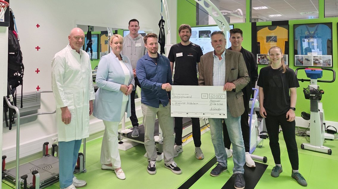 (von links): Uwe Tegtbur, Anke Mayer, Mirko Buzdon, Nico Kurpiers, Thorben Sundermeier, Wolfgang Schröter, Edward-Philipp Pieper, Lia Bernewitz stehen für ein Gruppenbild in einem Trainingsraum der Sportmedizin. Zwei von ihnen halten einen übergroßen Scheck in den Händen. 