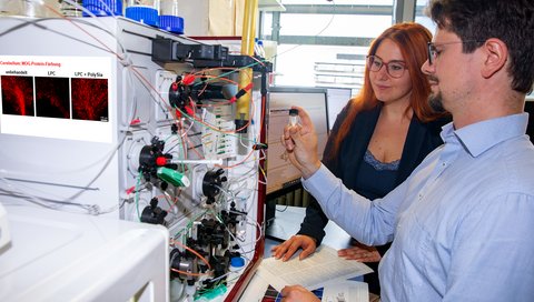 Dr Lara-Jasmin Schröder and Dr Hauke Thiesler stand in front of a chromatography device in a laboratory at the Institute of Clinical Biochemistry.