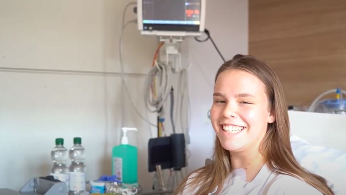 Patientin Annie sitzt auf dem Krankenbett und lächelt in die Kamera.